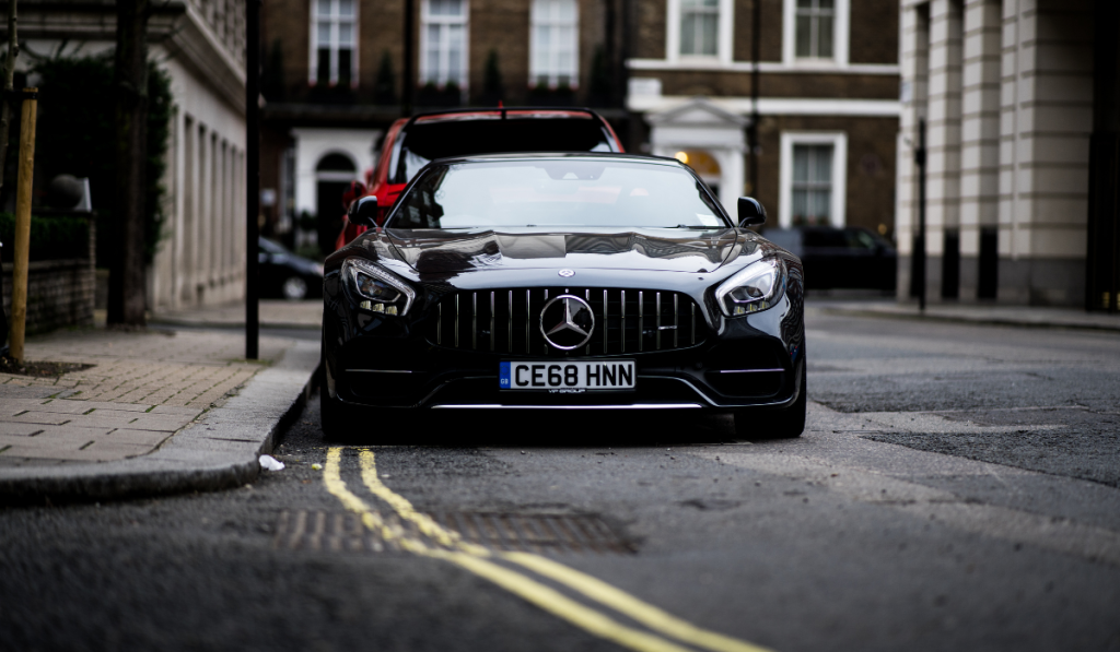 A black Mercedes-Benz sports car is parked on a city street, facing the camera. The license plate reads "CE68 HNN" and buildings are visible in the background.