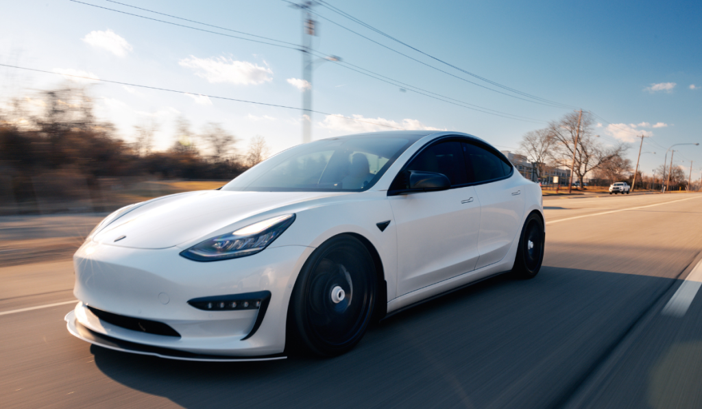 A white Tesla electric car drives down a sunny, empty road with blurred trees and power lines in the background, suggesting speed and motion.