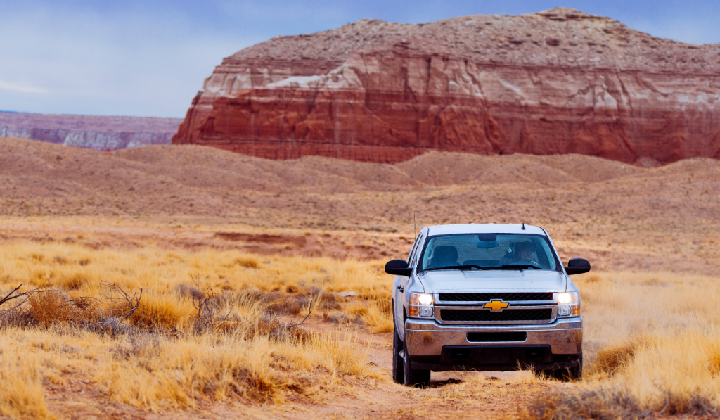 A white Chevrolet pickup truck drives on a dirt path through a dry, grassy desert landscape with a large red rock formation in the background under a cloudy sky.
