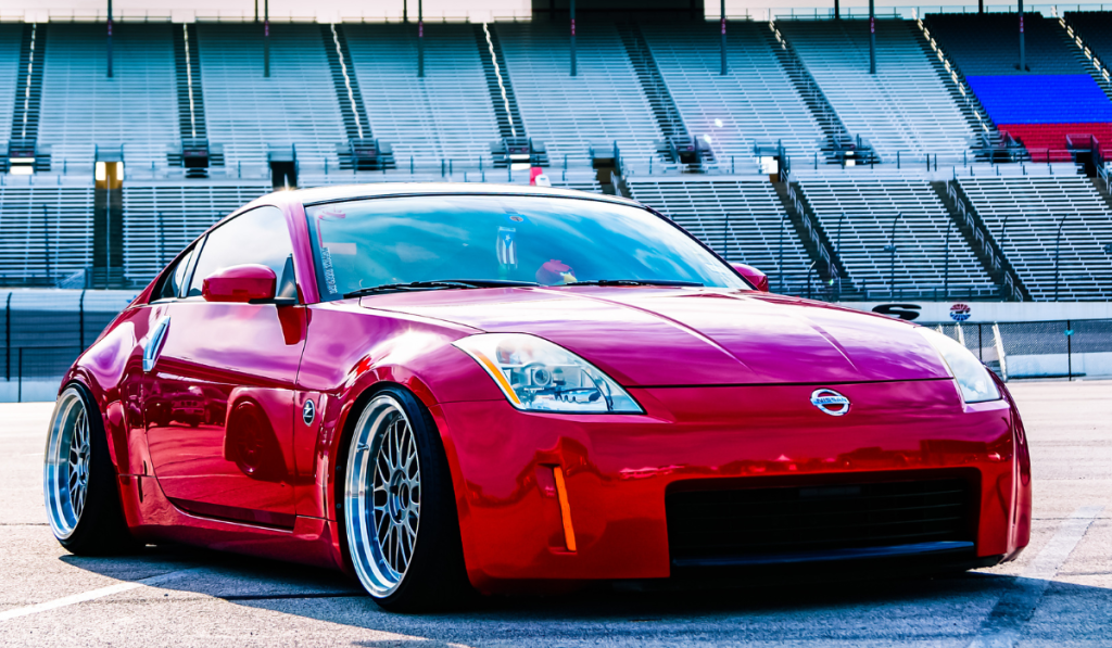 A shiny red Nissan 350Z sports car with custom wheels is parked on a race track in front of empty, tiered grandstands in daylight.