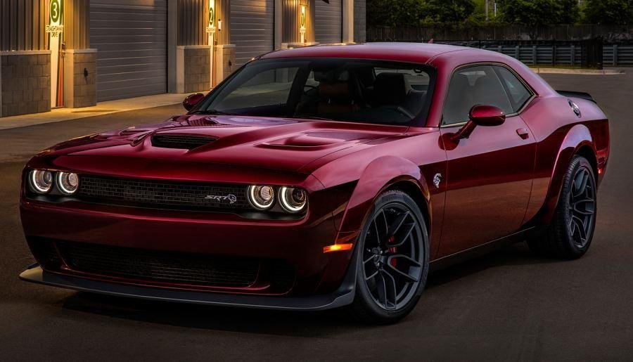 A red Dodge Challenger SRT is parked outdoors at dusk, with lit buildings in the background.