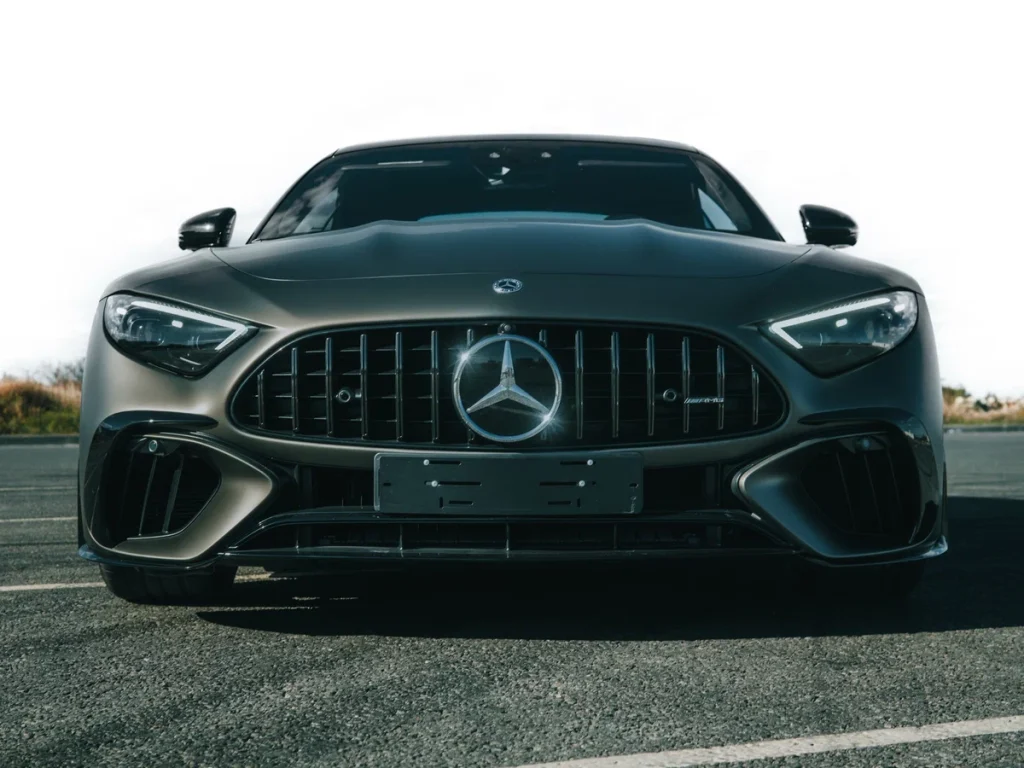 Front view of a black Mercedes-Benz car parked on pavement, with a prominent grille, shiny emblem, and modern headlights visible against a bright sky.