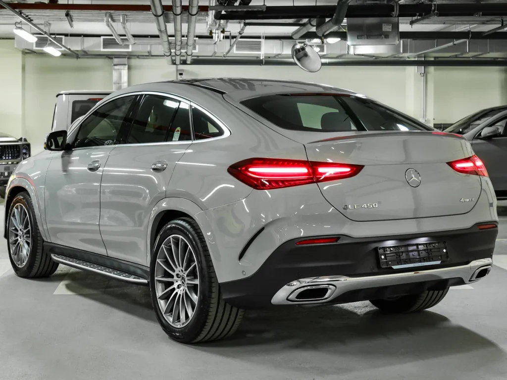 A silver Mercedes-Benz GLE 450 4MATIC SUV is parked indoors in a well-lit garage, viewed from the rear left angle, showcasing its sleek design and distinctive taillights.