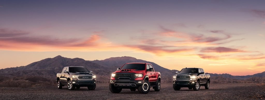 Three pickup trucks—black, red, and dark blue—are parked side by side on a rocky terrain, with mountains and a dramatic sunset sky in the background.