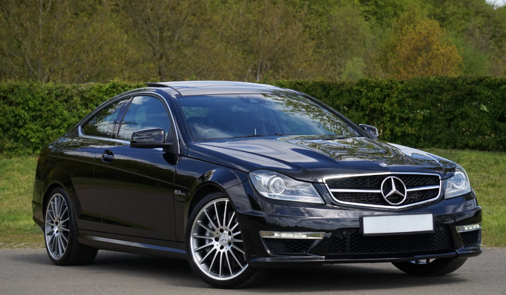 A sleek black Mercedes-Benz coupe parked on a road, with silver alloy wheels and a shiny finish. Green trees and bushes are visible in the background.