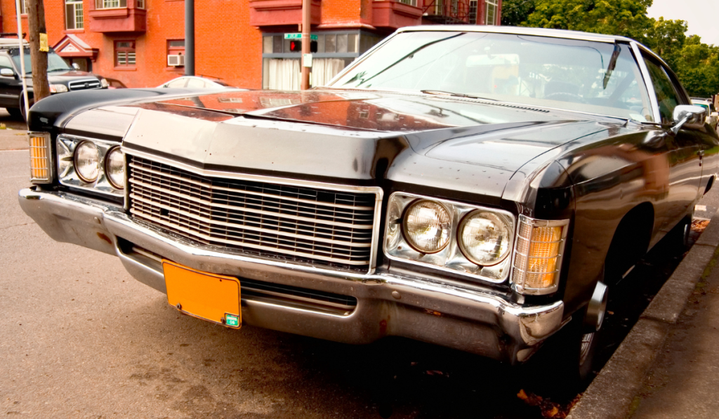 A close-up view of a vintage black car parked on a street beside a red brick building, showing its chrome grille, headlights, and yellow license plate.