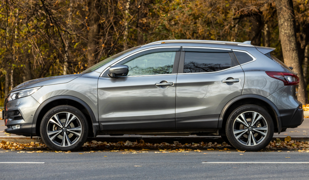 A silver SUV parked on the side of a road with autumn leaves scattered on the ground and trees with brown foliage in the background.