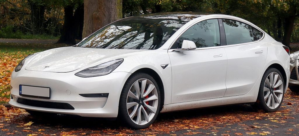 A white Tesla Model 3 electric car is parked on a street covered with fallen autumn leaves, with trees and greenery in the background.