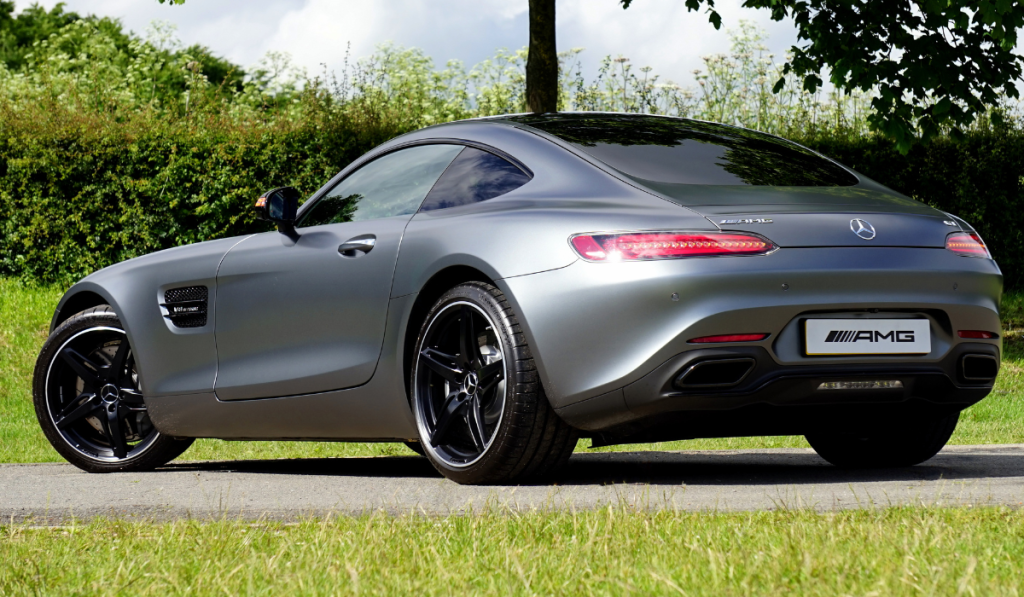 A sleek, silver Mercedes-Benz AMG sports car is parked on a road near green grass and bushes, with trees and a partly cloudy sky in the background. The car is viewed from the rear side.