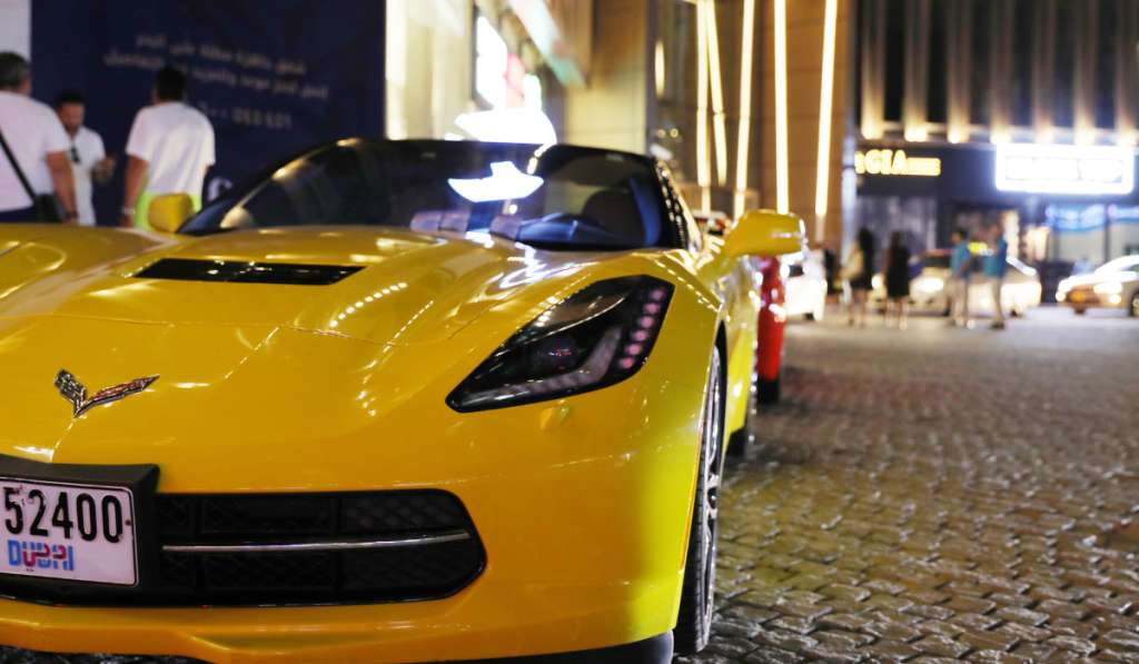 A bright yellow sports car is parked on a city street at night, with people and illuminated buildings visible in the background. The car's license plate features Arabic text and numerals.