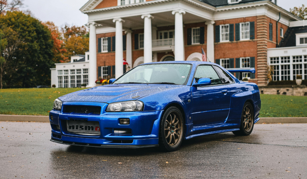 A blue Nissan Skyline sports car is parked on a wet driveway in front of a large, red-brick mansion with white columns and green lawns. The car is shiny and the scene appears to be on a rainy day.