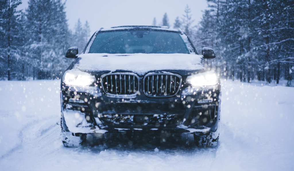 A black car covered in snow is parked on a snowy road in a forest, its headlights on during heavy snowfall. Snow blankets the ground and trees, emphasizing the importance of chandler windshield replacement for winter safety.