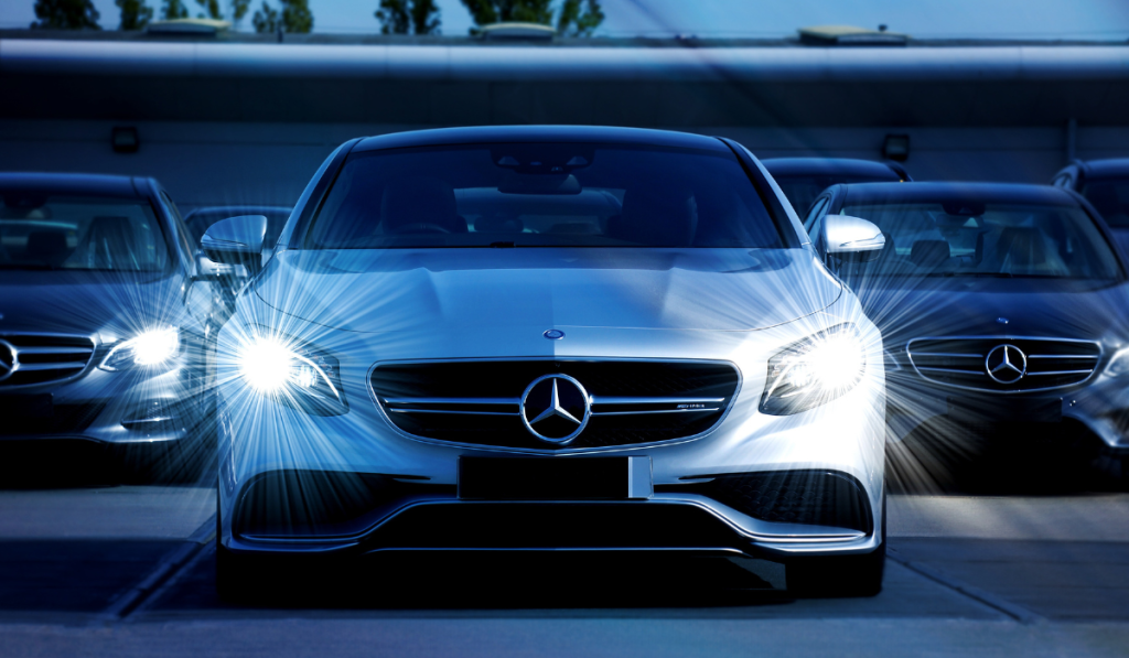 A silver Mercedes-Benz car with headlights on is parked in the foreground, flanked by two similar cars in a lot, with a blurred building and trees in the background.