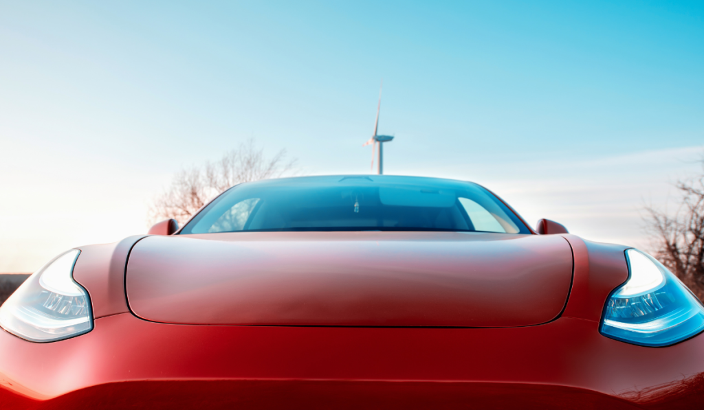A red electric car with headlights on is viewed from the front, parked outdoors with a wind turbine and bare trees visible in the background under a clear sky.