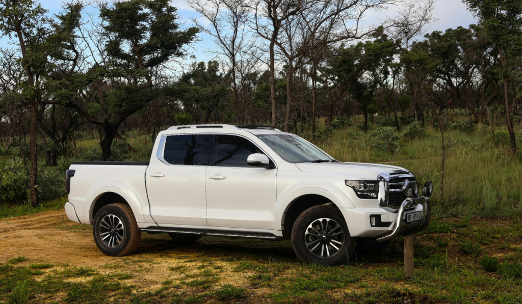 A white pickup truck is parked on a dirt path in a grassy, wooded area with trees and shrubs in the background under a partly cloudy sky.