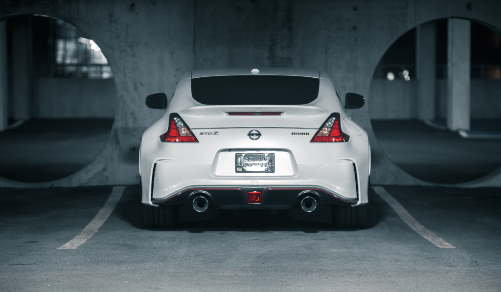 A white Nissan 370Z Nismo sports car is parked in a dimly lit concrete garage, viewed from the rear, with two large circular openings in the wall behind it.