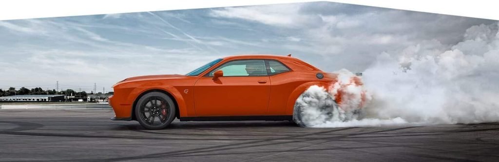 An orange muscle car performs a burnout on a racetrack, smoke pouring from rear tires.