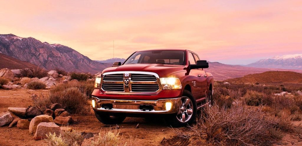 A red pickup truck is parked on a rocky, desert landscape with mountains in the background under a pink and orange sunset sky.