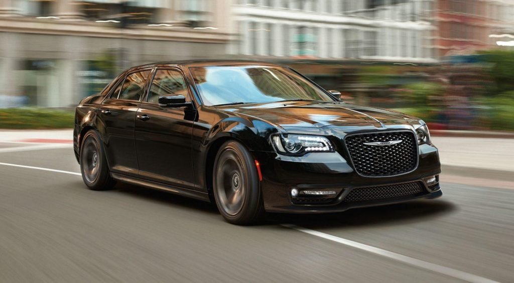 A black Chrysler 300 sedan speeds down a city street, passing blurred buildings in the urban background.