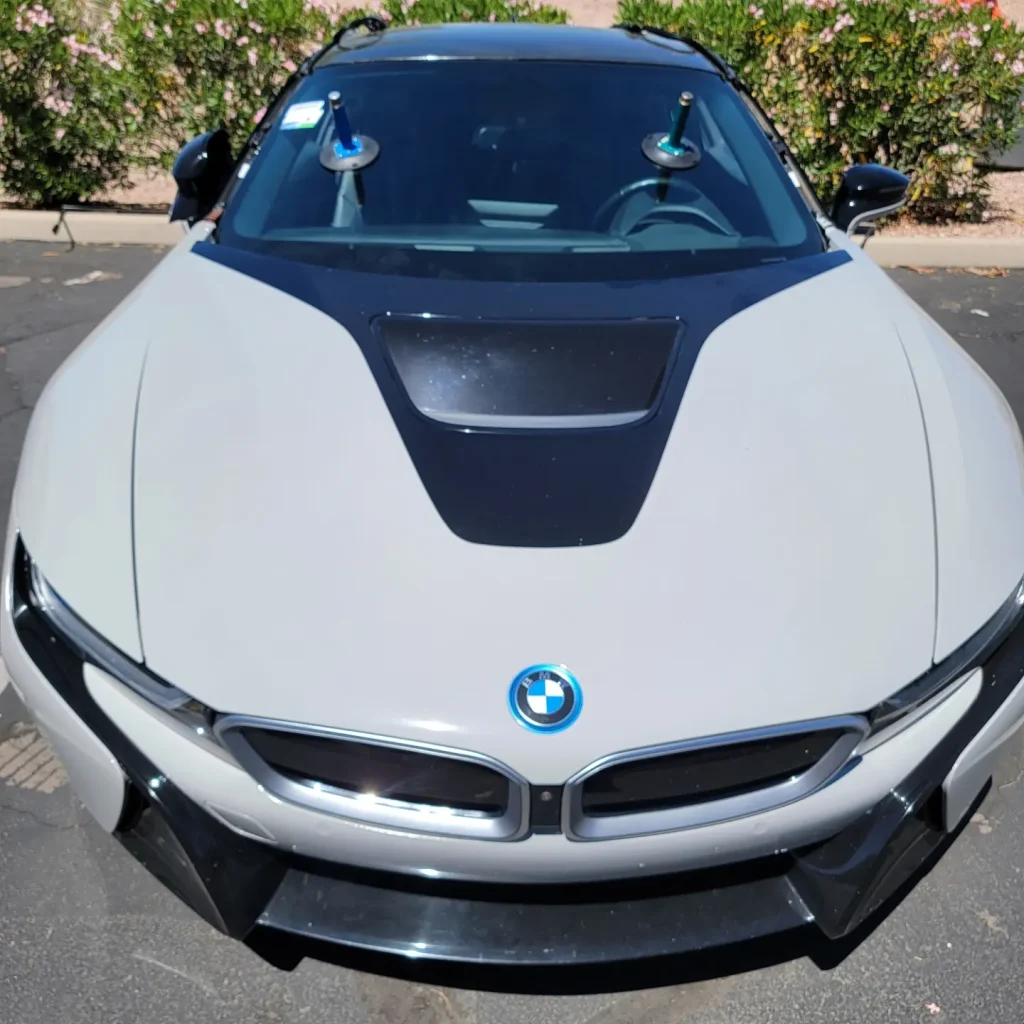 A front view of a silver BMW sports car parked outdoors, its windshield featuring two suction-cup-mounted devices, possibly for chandler windshield replacement. Bushes and a building appear in the background.