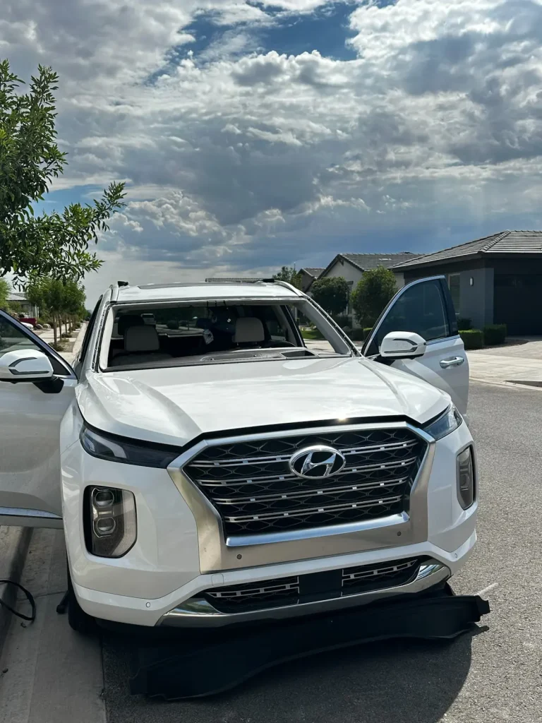 A white Hyundai SUV is parked on a suburban street with both front doors open. The sky is cloudy, and a black mat or cover is laid out in front of the vehicle on the road. Houses and trees are visible in the background.