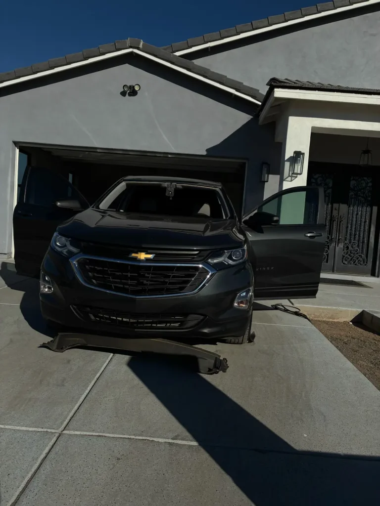 A black Chevrolet SUV is parked in a driveway in front of a gray house, with its front doors and sunroof open. The car’s front wheels are missing, and it sits on jacks.