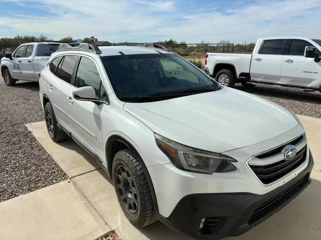 A white Subaru Outback is parked on a concrete driveway next to a gravel lot, with a white pickup truck parked nearby under a partly cloudy sky.