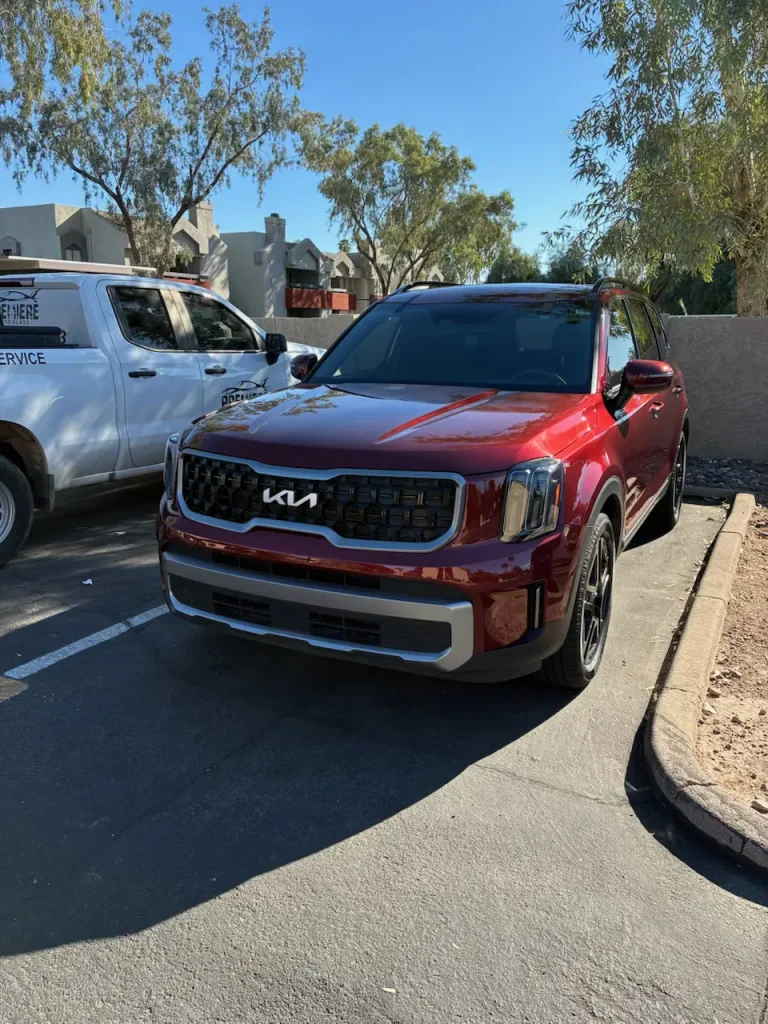 A red Kia SUV is parked in a sunny parking lot next to a white service truck, with trees and buildings in the background.