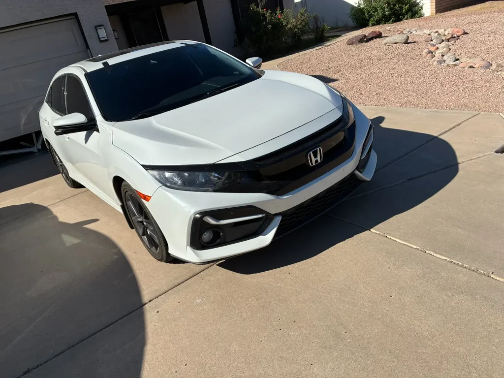 A white Honda Civic hatchback is parked on a concrete driveway next to a gravel yard and a house on a sunny day.