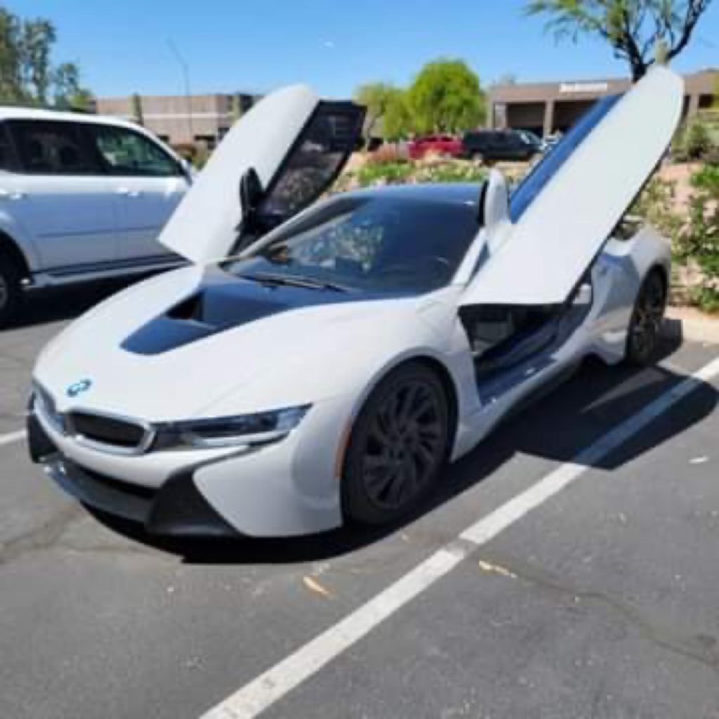 A white BMW i8 sports car with its butterfly doors open is parked in a sunny parking lot next to a white SUV, perfect for those seeking Chandler windshield replacement amid trees and buildings in the background.