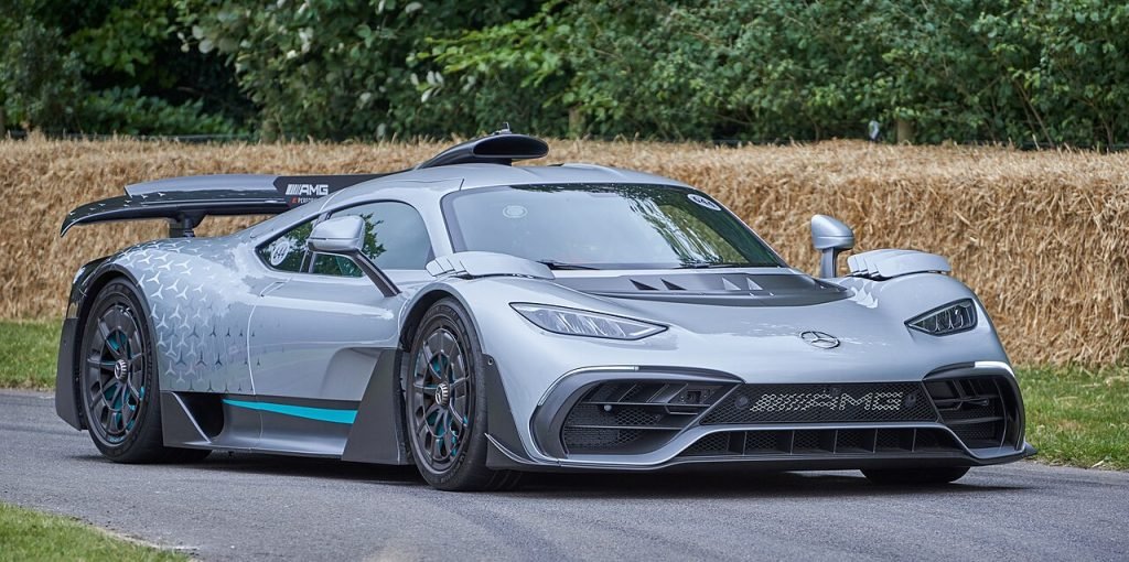 A silver Mercedes-AMG One hypercar with aerodynamic design and black accents is parked on a paved road, with green foliage and a straw barrier in the background.