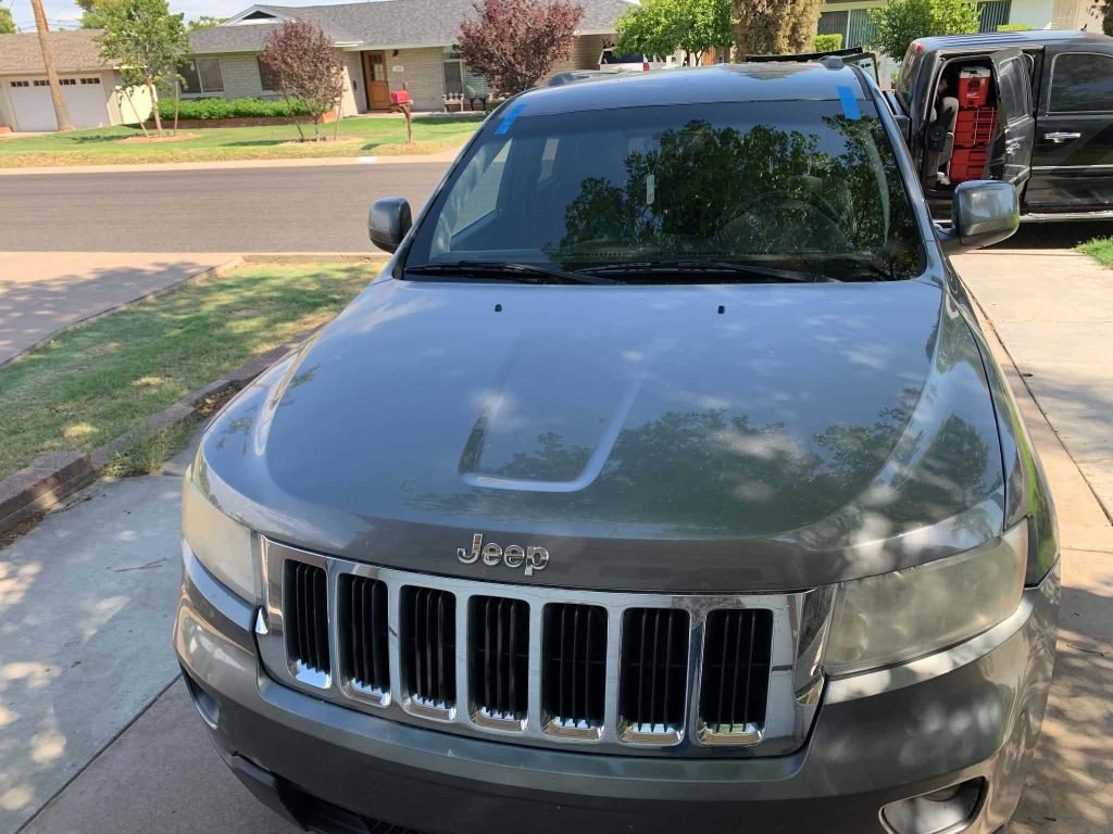 A gray Jeep SUV is parked in a driveway on a sunny day, with trees and houses visible across the street in a suburban neighborhood.