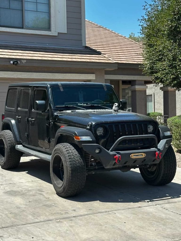 A black Jeep Wrangler with large tires is parked in a driveway in front of a suburban home.
