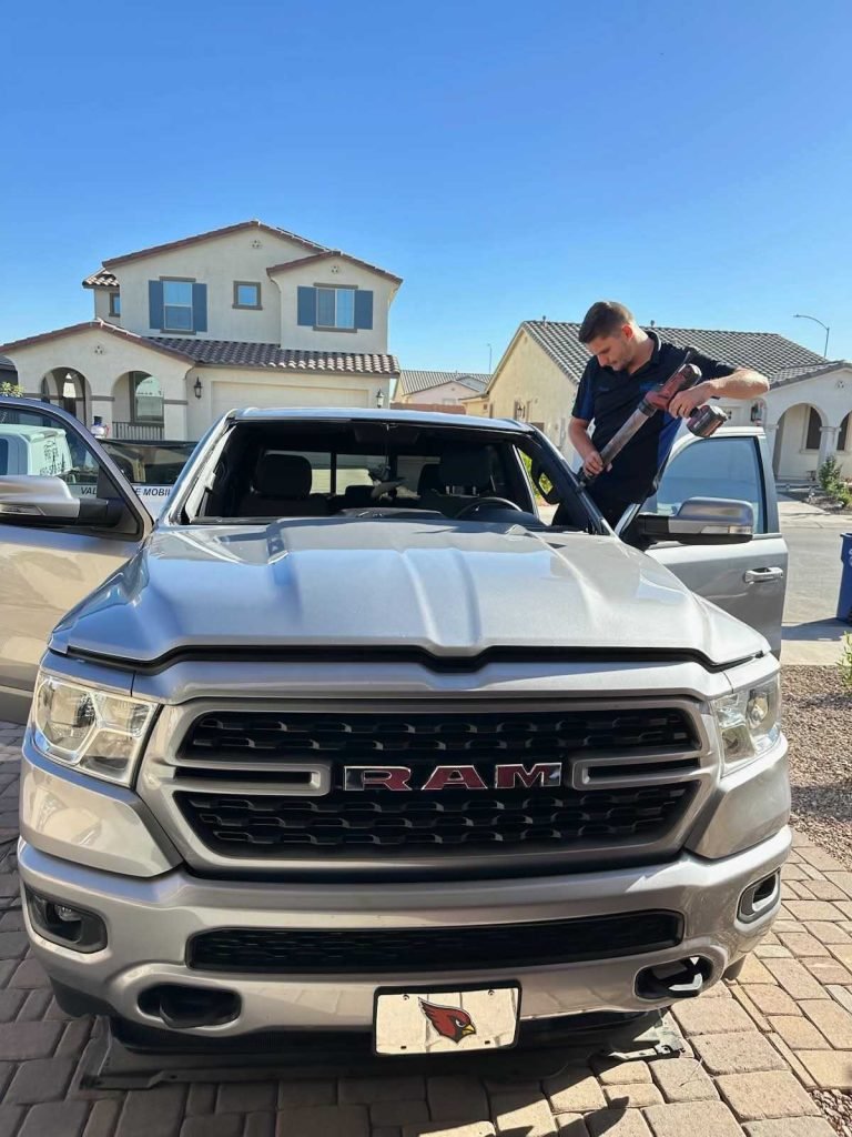 A man stands on the passenger side of a silver RAM truck, working on the windshield with tools. The truck is parked in a residential driveway with houses and a clear blue sky in the background.