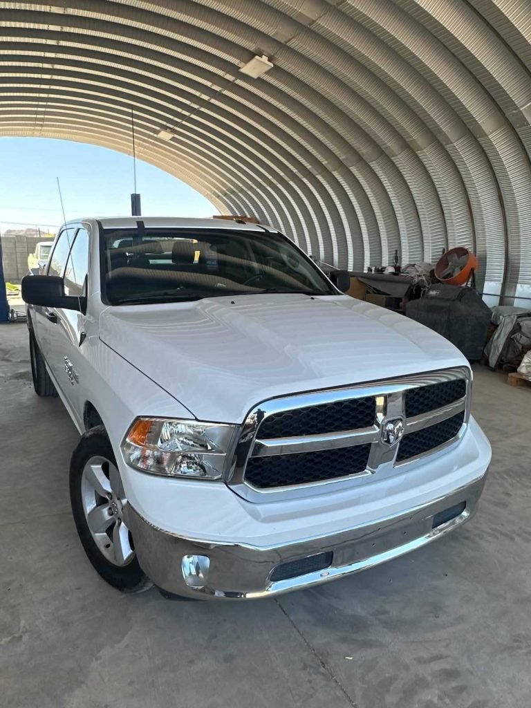 White Dodge Ram pickup truck parked in a curved metal garage, various items loaded in the truck bed.