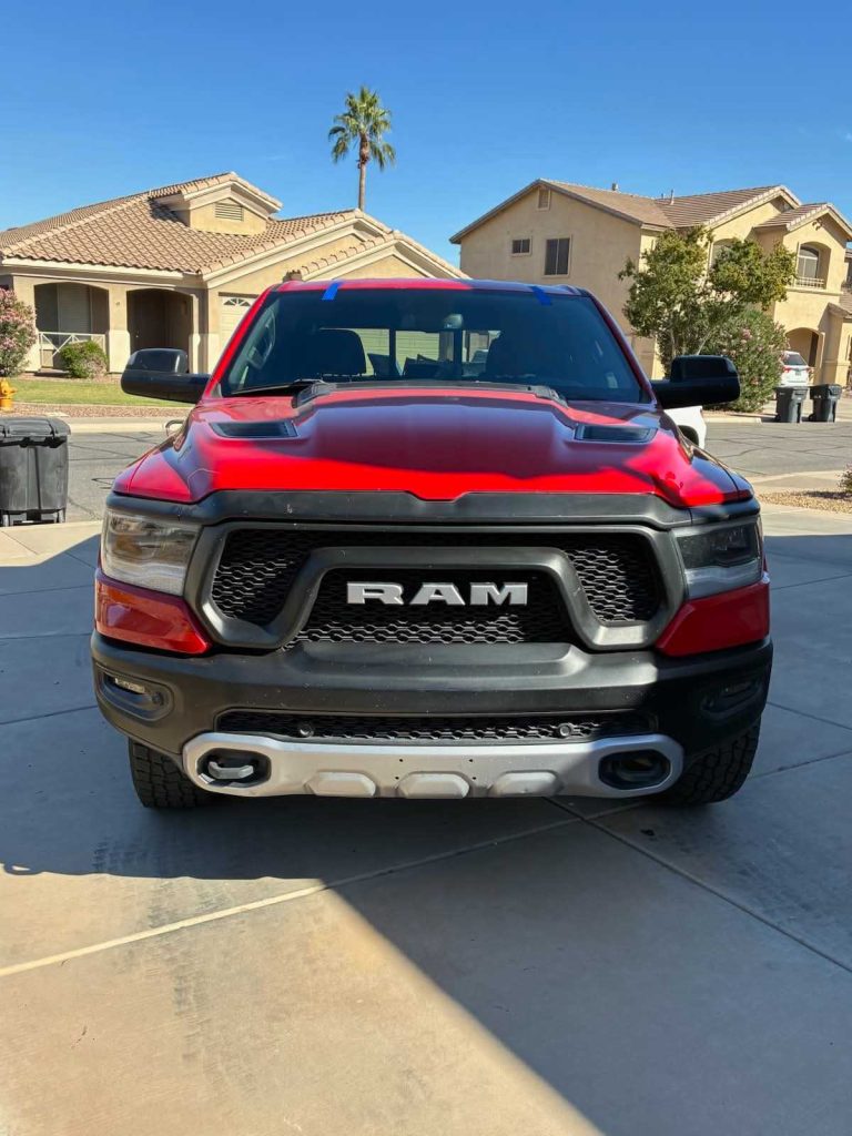 A red RAM pickup truck is parked on a driveway in front of suburban houses on a sunny day, with a clear blue sky and a tall palm tree in the background.