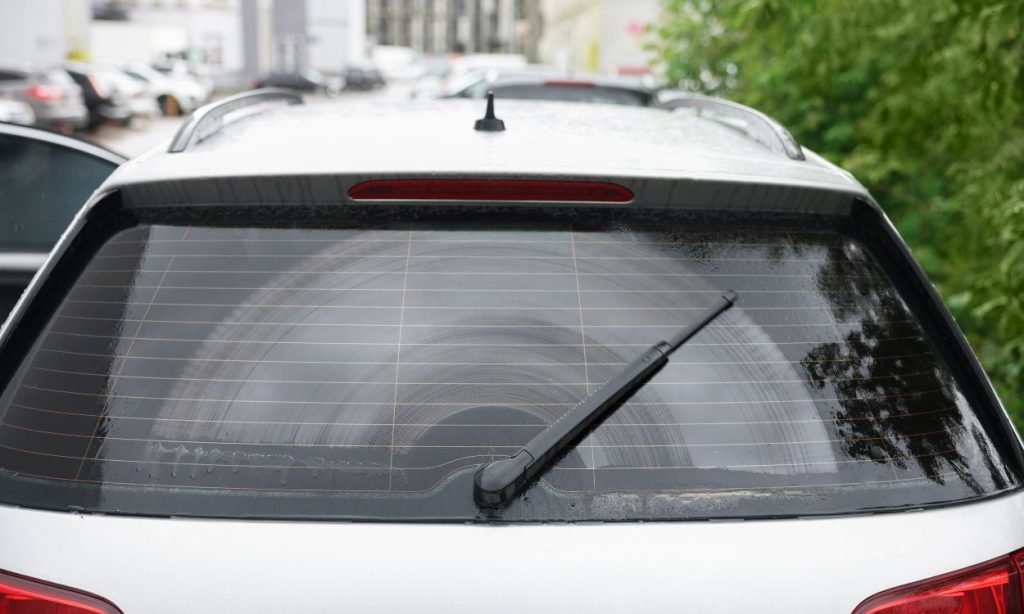 A rear view of a car with its back windshield partially cleaned by the rear wiper, leaving a clear semi-circular area on the otherwise wet and foggy glass. Trees and other cars are visible in the background.