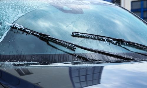 A close-up of a car windshield with water on it as two windshield wipers move across the glass, clearing away the water. Reflections of nearby buildings are visible on the windshield.