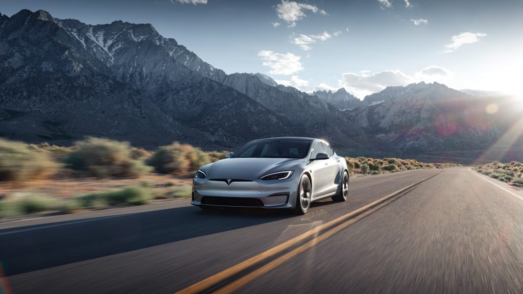 A silver Tesla car drives on an open highway through a scenic mountainous landscape under a partly cloudy sky, with sunlight shining from the right side of the image.