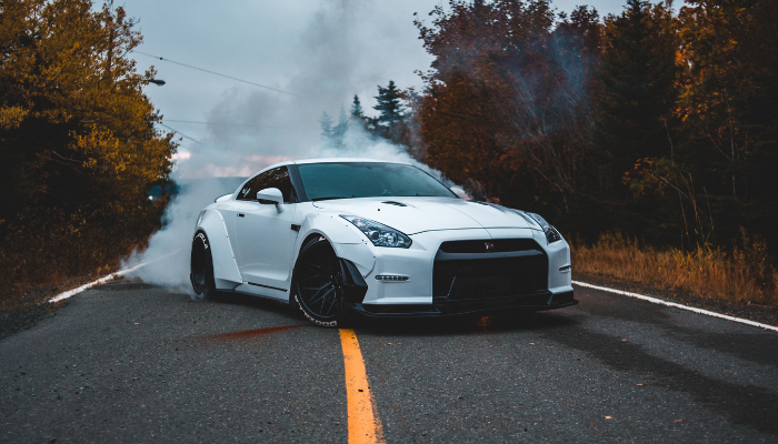 A white sports car does a burnout on a forest-lined road, creating clouds of smoke around its tires. The car is angled slightly, and the scene has an autumn feel with colorful leaves on the trees.