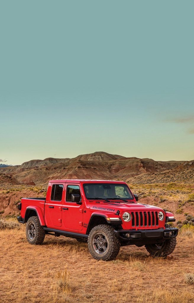 A bright red Jeep pickup truck is parked on dry grass in a rugged, hilly desert landscape under a clear sky with a wide expanse of blue above.