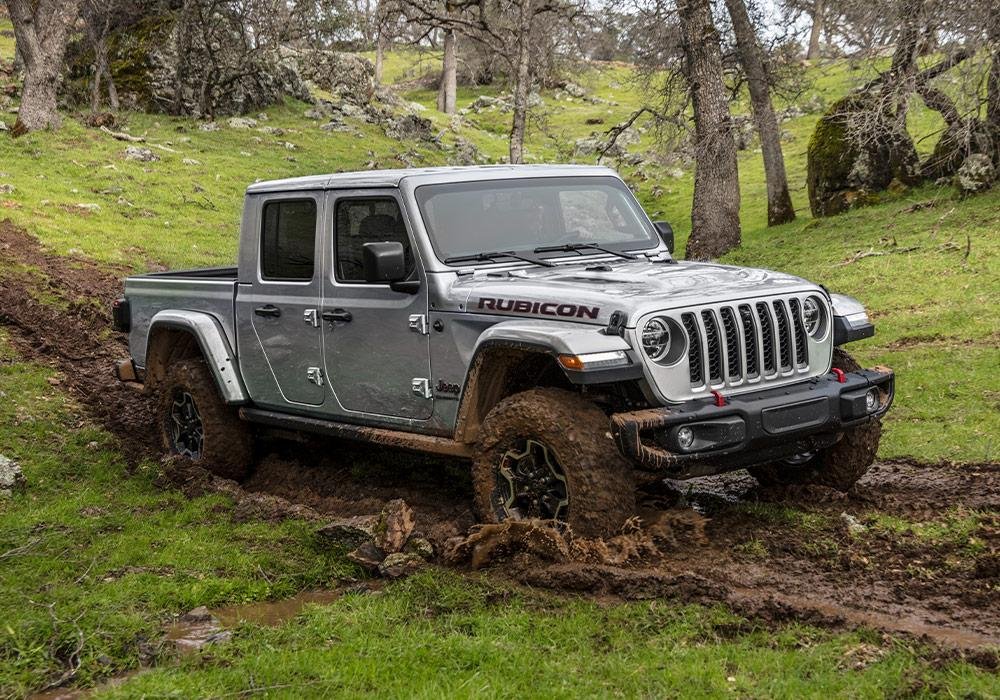 A silver Jeep Gladiator Rubicon pickup truck drives through muddy terrain in a grassy, wooded area, with mud splashed on its wheels and lower body.