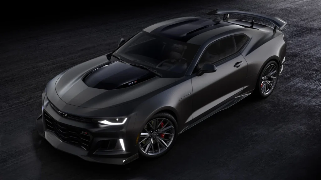 A sleek, black Chevrolet Camaro sports car is parked on a dark surface, viewed from above at an angle, highlighting its aerodynamic design, aggressive front grille, and prominent rear spoiler.