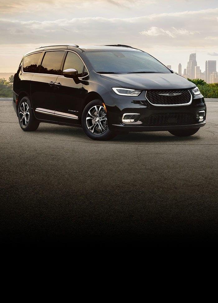 A black minivan is parked on a road, with a city skyline and a cloudy sky visible in the background.