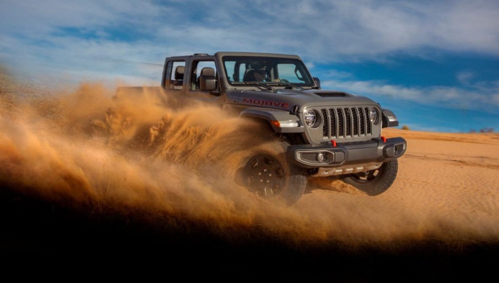 A gray Jeep Gladiator Mojave kicks up sand as it speeds over a dune in a desert landscape under a partly cloudy blue sky.