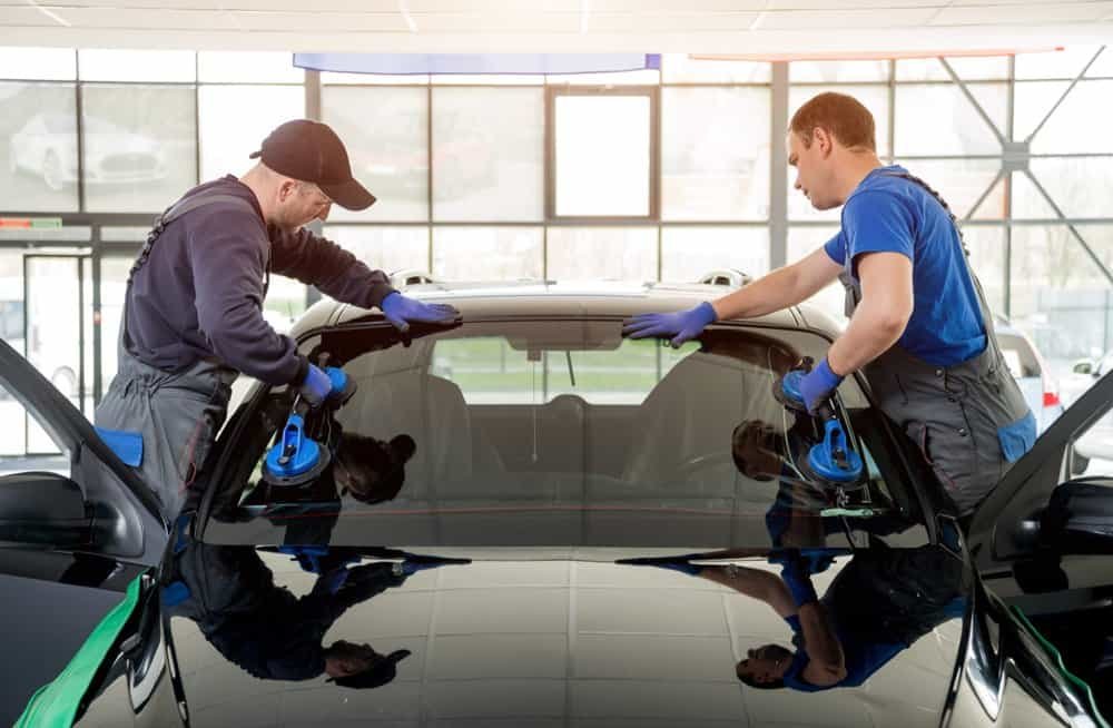 Two workers in overalls and gloves are installing a windshield on a black car using suction cups. With precision akin to ADAS calibration, they are focused on aligning the glass in a well-lit indoor setting with large windows in the background.