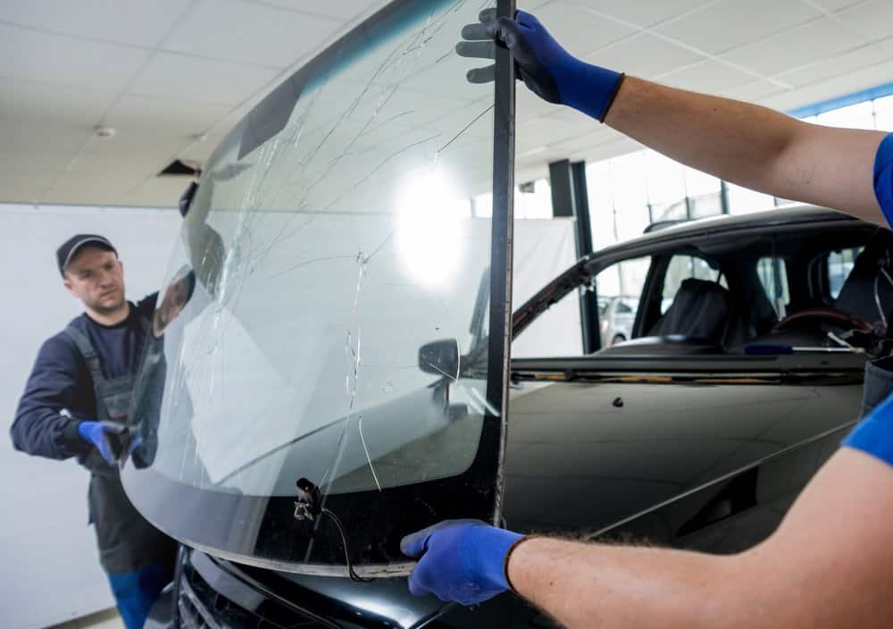 Two auto technicians in a well-lit garage are carefully performing a windshield repair, skillfully removing a cracked windshield from a car. They are wearing blue gloves and working together to lift the damaged glass away from the vehicle with precision.