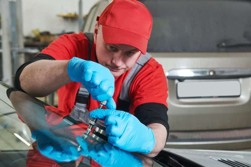 A mechanic in a red cap and overalls, wearing blue gloves, expertly performs a windshield repair on a beige car using a specialized tool. The vehicle is parked in a garage setting.