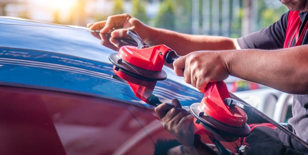 A person carefully uses a red glass suction lifter to handle a large glass sheet, reminiscent of windshield replacement work. They wear a red and black uniform, standing in a brightly lit scene with greenery softly blurred in the background.