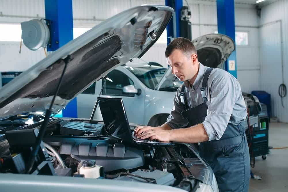 A mechanic in a workshop wearing overalls is using a laptop to diagnose a car engine. The car's hood is open, and another vehicle is visible in the background. Blue pillars and tools for windshield replacement surround the area.